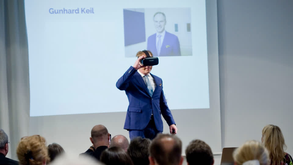 Gunhard Keil auf der Bühne setzt eine VR-Brille auf, vor ihm ein voller Saal