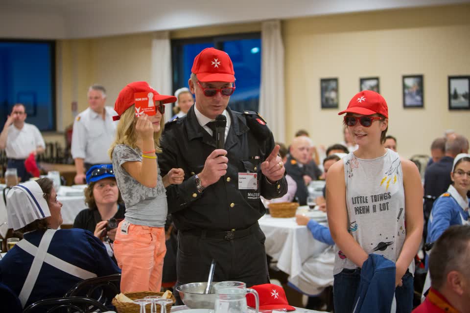 Gunhard Keil in Malteser-Uniform mit roter Kappe und Sonnenbrille spricht ins Mikrofon, neben ihm zwei Mädchen mit roten Kappen, dahinter ein voller Speisesaal