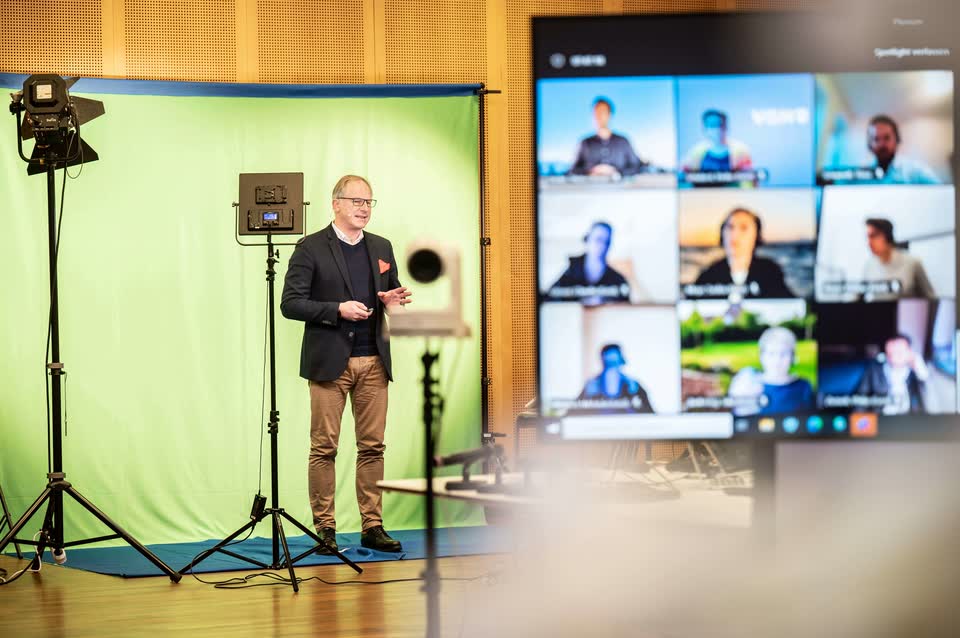 Gunhard Keil spricht im Studio vor einer Greenscreen-Wand, rechts ein Bildschirm mit den Online-Teilnehmenden