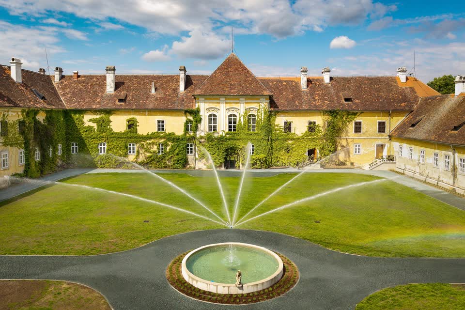 Der Innenhof von Schloss Kohfidisch von oben: gelbe Fassaden mit wildem Wein, frischer Rasen, fächerförmige Wasserstrahlen der Bewässerung und ein runder Brunnen